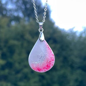 May include: A silver chain necklace with a teardrop pendant. The pendant is white with a pink and silver glitter design. The pendant has a silver initial "G" in the center.