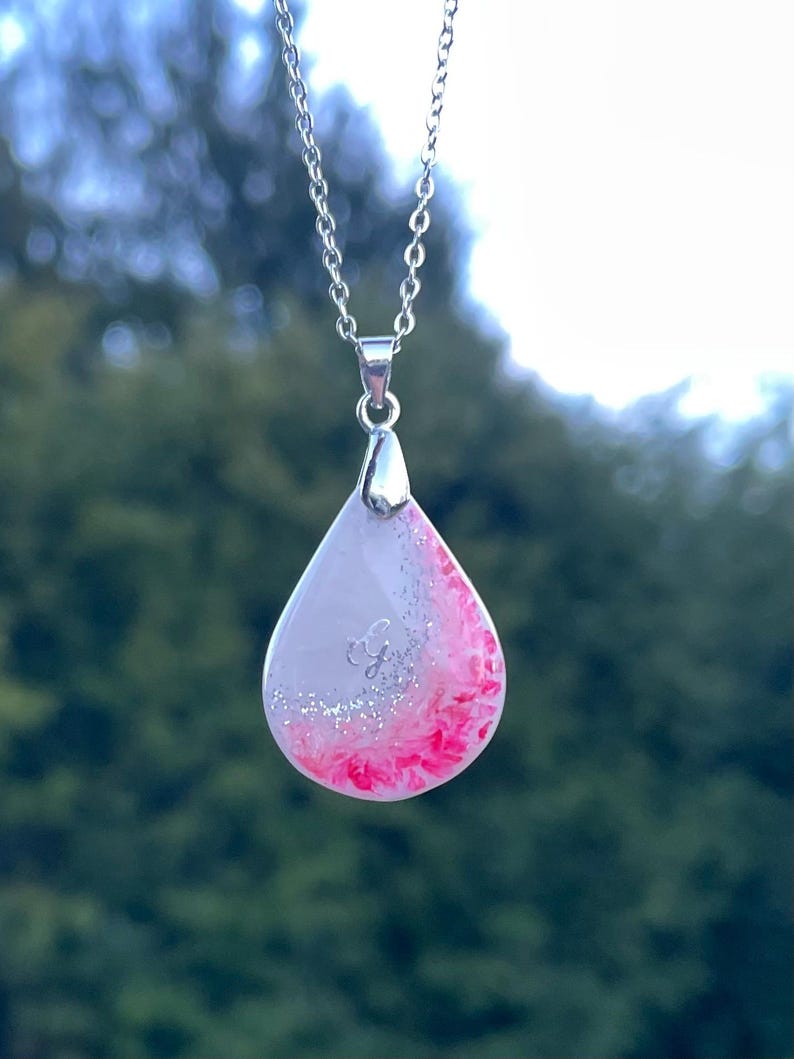 May include: A silver chain necklace with a teardrop pendant. The pendant is white with a pink and silver glitter design. The pendant has a silver initial "G" in the center.