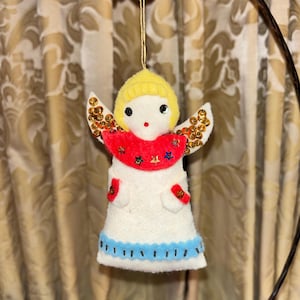 May include: A handmade Christmas angel ornament. The angel has a yellow head, white body, and red accents. The wings are embellished with gold sequins. The angel's dress has a blue scalloped hemline. The ornament is suspended by a gold string.