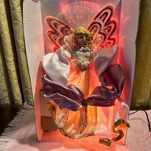May include: A decorative angel figurine with a blonde head and a flowing gown. The angel has large, ornate wings and is surrounded by fiber optic lights. The figurine is set inside a pink box with a pink bow.