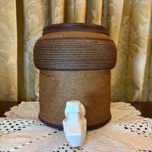 May include: A large, brown ceramic water cooler with a white spigot. The cooler has a textured, horizontal-lined design around the middle. The top and bottom rims are a darker brown. The cooler sits on a white, lace doily.