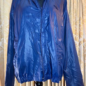 May include: A blue nylon jacket with a zip-up front, a stand-up collar, and two front pockets. The jacket has a small logo on the chest and elastic cuffs and hem. The jacket is displayed on a black mannequin.