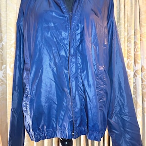 May include: A shiny blue windbreaker jacket featuring a high collar with snap button closures, a full-length front zipper, and long sleeves with elastic cuffs. The jacket has a smooth, reflective finish and a simple, functional design.