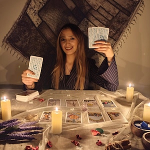 May include: A set of tarot cards with intricate blue and white patterns on the back, held by a person in a dark pinstriped blazer. The table is covered with a light cloth, scattered tarot cards, lit pillar candles, dried lavender, and cinnamon.