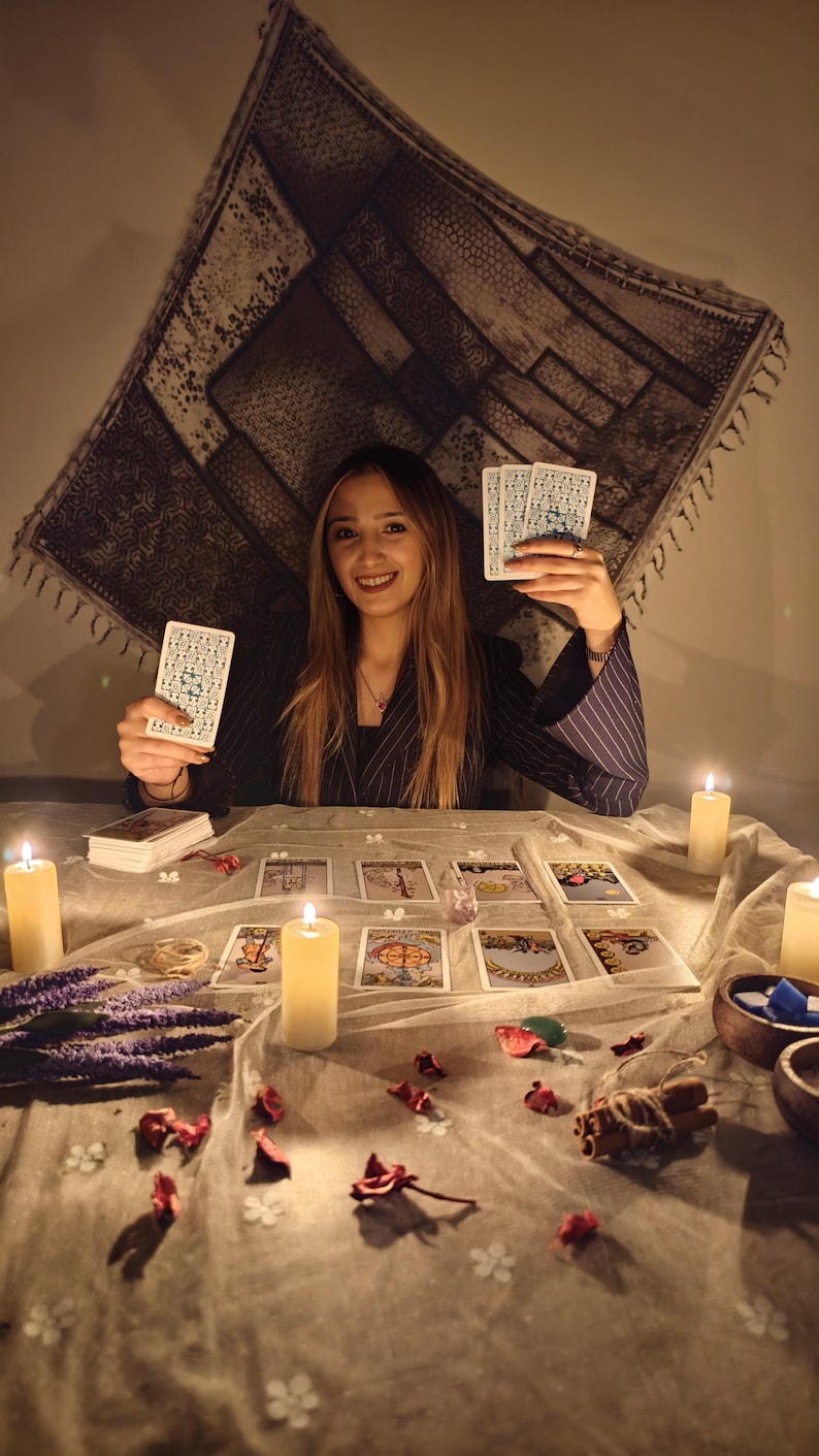 May include: A set of tarot cards with intricate blue and white patterns on the back, held by a person in a dark pinstriped blazer. The table is covered with a light cloth, scattered tarot cards, lit pillar candles, dried lavender, and cinnamon.
