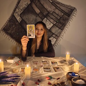 May include: A tarot card reading setup featuring a deck of cards, with one card labeled The Chariot held up. The table is covered with a light cloth, scattered dried flower petals, cinnamon sticks, small wooden bowls, and several lit pillar candles.