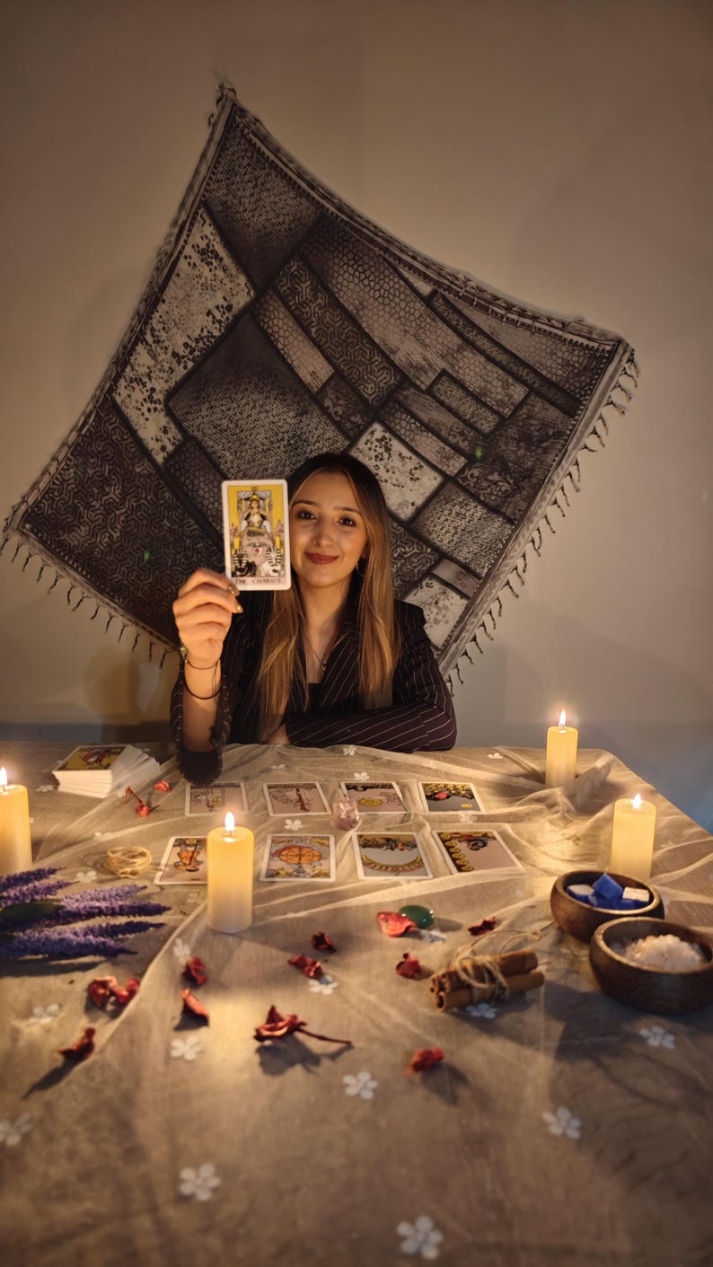 May include: A tarot card reading setup featuring a deck of cards, with one card labeled The Chariot held up. The table is covered with a light cloth, scattered dried flower petals, cinnamon sticks, small wooden bowls, and several lit pillar candles.