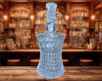 May include: A clear, cut-glass decanter with a diamond pattern on the lower portion and a decorative stopper. The decanter is displayed on a wooden surface, with a blurred bar scene in the background.