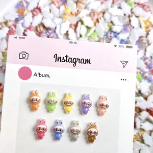 May include: A pink Instagram album display featuring eleven colorful bunny figurines with eerie smiles. The bunnies are in various colors, including orange, green, yellow, purple, pink, blue, gray, and brown. The background is filled with similar figurines.
