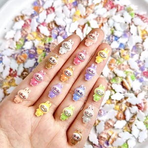 May include: A collection of colorful, small bunny-shaped charms in various colors, including pink, yellow, and blue. Each charm has a smiling face and is held in a hand, with more charms scattered in the background. These charms are likely for crafting or jewelry making.