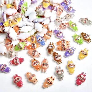May include: A collection of colorful, small bunny-shaped charms in various colors, including red, orange, yellow, green, blue, purple, and gray. The charms have a smiling face design and are scattered across a white surface.