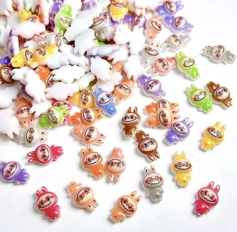 May include: A collection of colorful, small bunny-shaped charms in various colors, including red, orange, yellow, green, blue, purple, and gray. The charms have a smiling face design and are scattered across a white surface.