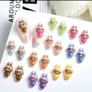 May include: A collection of colorful bunny-shaped nail art decorations. The bunnies come in various colors, including brown, pink, green, orange, blue, yellow, gray, and purple. Some are applied to painted nail tips. The bunnies have a cartoonish design with a smiling face.