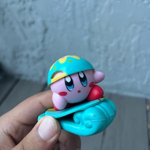 May include: A small, pink Kirby figurine with blue eyes and a red mouth, wearing a teal helmet with yellow accents, riding a teal skateboard. The figurine is held in a hand against a blurred background.