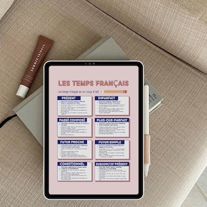 May include: A tablet showing French grammar, with the title "LES TEMPS FRANÇAIS." A tube of Summer Fridays skincare and a pen are also visible. The tablet rests on a book, all on a beige surface.