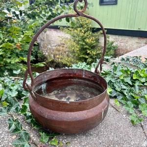 May include: A vintage copper pot with a rustic iron handle. The pot has a rounded bottom and a wide opening. The pot is slightly tarnished, giving it a weathered look.