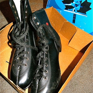 May include: A pair of black leather ice skates with black laces and a white lining. The skates are in a cardboard box with a blue and white maple leaf design.