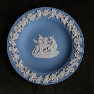 May include: A blue and white ceramic plate with a white relief design of a scene with three figures. The plate has a white border with a floral design.