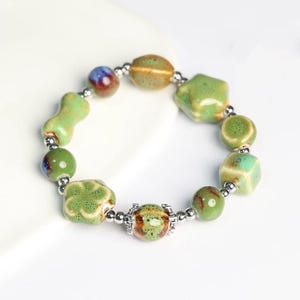 May include: A green ceramic bracelet with various shaped beads, including stars, flowers, and squares. The beads are connected with silver spacers.