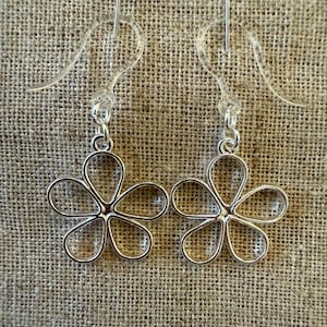 May include: A pair of silver flower-shaped dangle earrings with a simple design. The earrings are on a brown fabric background.