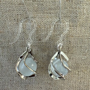 May include: A pair of silver earrings with a clear glass orb suspended in a cage-like design. The earrings have clear plastic hooks.