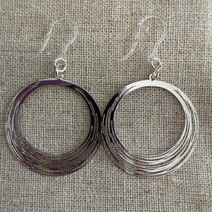 May include: A pair of silver-toned hoop earrings. Each earring features multiple concentric circles, creating a layered effect. The earrings hang from clear, plastic ear wires. The background is a textured, neutral-colored fabric.