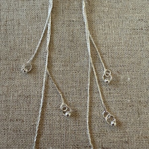 May include: Silver-toned dangle earrings with multiple strands. Each strand features a small silver bead and a delicate loop design. The earrings are displayed against a neutral, textured background.
