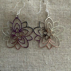 May include: A pair of silver-colored flower-shaped earrings with clear plastic ear wires. The earrings have a layered design, with each layer forming a petal. The earrings are displayed on a textured, neutral-colored surface.