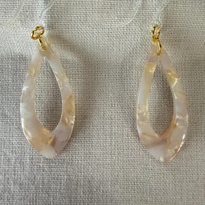 May include: A pair of teardrop-shaped earrings. The earrings have a pearlescent, off-white color with subtle beige marbling. The earring hooks are gold-colored. The earrings are hanging against a neutral fabric background.