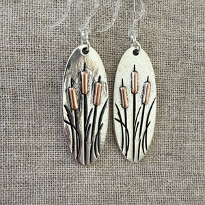 May include: A pair of silver oval earrings with a copper design of cattails. The earrings have a hook closure.