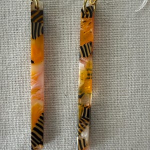 May include: Two rectangular earrings with gold-colored hooks. The earrings are primarily orange with black and white striped accents. The earrings are suspended against a neutral, textured background.