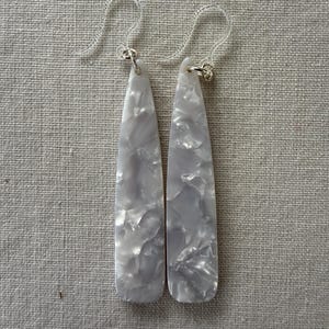 May include: A pair of long, teardrop-shaped earrings featuring a white and light gray marbled pattern. The earrings are suspended from clear plastic ear hooks attached with small silver-toned metal jump rings, creating a sleek, elongated look.