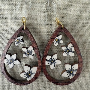 May include: Wooden teardrop earrings with a dark brown frame and white flower cutouts. The earrings have gold-colored hooks. The flowers have black and yellow centers. The earrings are displayed on a tan fabric background.