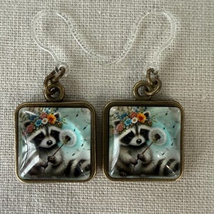May include: A pair of square earrings with a whimsical raccoon design. Each earring features a raccoon wearing a floral crown and holding a dandelion. The earrings have a bronze-colored frame and clear ear wires.