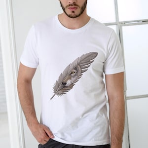 May include: White t-shirt featuring a detailed illustration of a wolf's face within a feather design. The wolf's eye is yellow, and the feather is a mix of gray and brown. The shirt is a classic crew neck style.