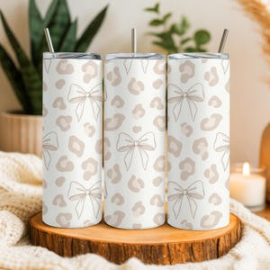 May include: Three white stainless steel tumblers with a bow and leopard print design. Each tumbler has a silver straw and lid. The tumblers are displayed on a wooden slice.