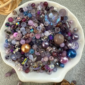 May include: A collection of assorted beads in a white seashell-shaped dish. The beads are in shades of purple, pink, and blue, with various shapes and sizes, including pearls, faceted beads, and round beads. Some beads have an iridescent finish, and a large, clear, flower-shaped bead is visible.