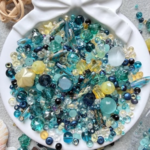 May include: A collection of assorted beads in shades of teal, turquoise, navy blue, and pale yellow, contained within a white shell-shaped dish. The beads vary in size, shape, and texture, including faceted, smooth, round, and star-shaped pieces.