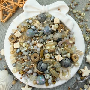May include: A white, bow-shaped dish filled with a variety of beads in shades of beige, gray, and iridescent colors. The beads include round, square, and irregular shapes, creating a textured and visually appealing display. A few seashells and a star-shaped object are also visible.