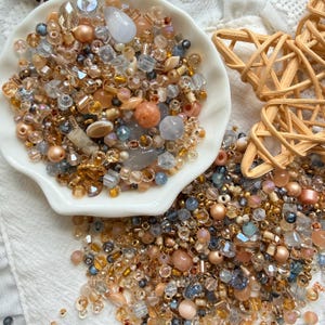 May include: A collection of assorted beads in shades of gold, peach, beige, and translucent gray, featuring various shapes including round, faceted, and cube forms. Some beads are contained within a white scalloped dish next to a wooden star.