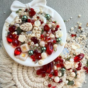 May include: A white, bow-shaped dish overflowing with a variety of beads. The beads are in shades of red, cream, green, and clear. Some beads are faceted, others are round, and some have leaf or star designs. The dish sits on a woven coaster.