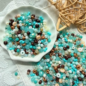 May include: A collection of assorted beads in shades of turquoise, brown, cream, and clear, displayed in a white seashell-shaped dish and scattered on a white surface. The beads vary in size and shape, suitable for jewelry making.