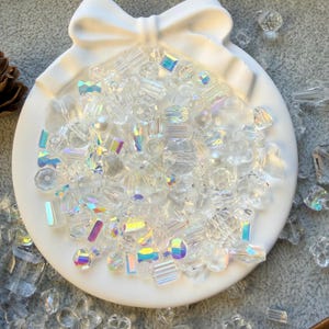 May include: A collection of clear, iridescent beads in various shapes and sizes, including faceted, round, and floral designs, are arranged on a white, bow-shaped dish. The beads reflect light, creating a rainbow effect. The dish is on a neutral background.