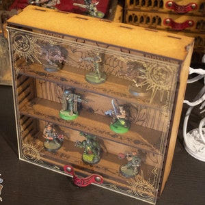 Puede incluir: Una vitrina de madera con estantes de vidrio para figuras en miniatura. La vitrina tiene un diseño dorado y negro con un borde decorativo. La vitrina está llena de figuras en miniatura, incluyendo una figura verde con una espada, una figura azul con una pistola y una figura roja con un casco.