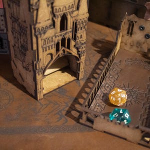 May include: A wooden tower with a dice rolling tray. The tower has a gothic design and is made of light brown wood. The tray is also made of wood and has a dark brown finish. There are two dice on the tray, one is gold and one is blue.