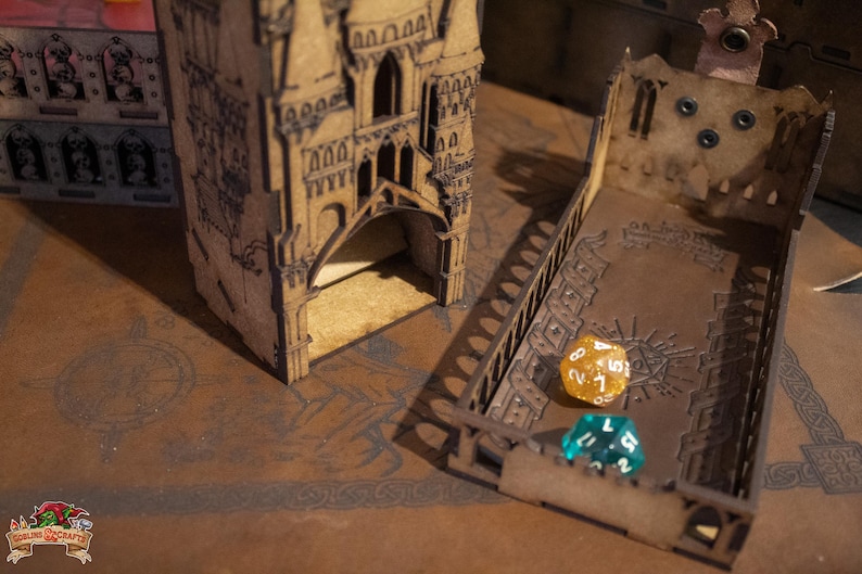 May include: A wooden tower with a dice rolling tray. The tower has a gothic design and is made of light brown wood. The tray is also made of wood and has a dark brown finish. There are two dice on the tray, one is gold and one is blue.