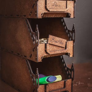 Könnte beinhalten: Ein dreistufiger brauner Holz-Organizer mit offenen Fächern. Das oberste Fach ist mit Pens beschriftet, das mittlere mit Buckles und das untere ist leer. Die Seitenwände weisen dekorative Ausschnitte auf, die an gotische Bögen erinnern.