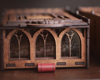 May include: A rectangular brown wooden box featuring three arched window cutouts with intricate tracery details. The box has a dark brown frame and a small, cylindrical red leather pull tab attached to the front center of the base structure.