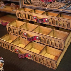 May include: A wooden box with three drawers, each with multiple compartments. The drawers have red leather pulls and intricate carvings. The box is made of light brown wood and has a dark brown stain.