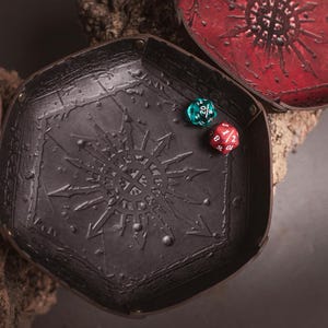 May include: A black leather dice tray with a sunburst design and two multi-sided dice. The tray is hexagonal and has a textured surface. A red and a teal die are inside the tray. A red tray is in the background.